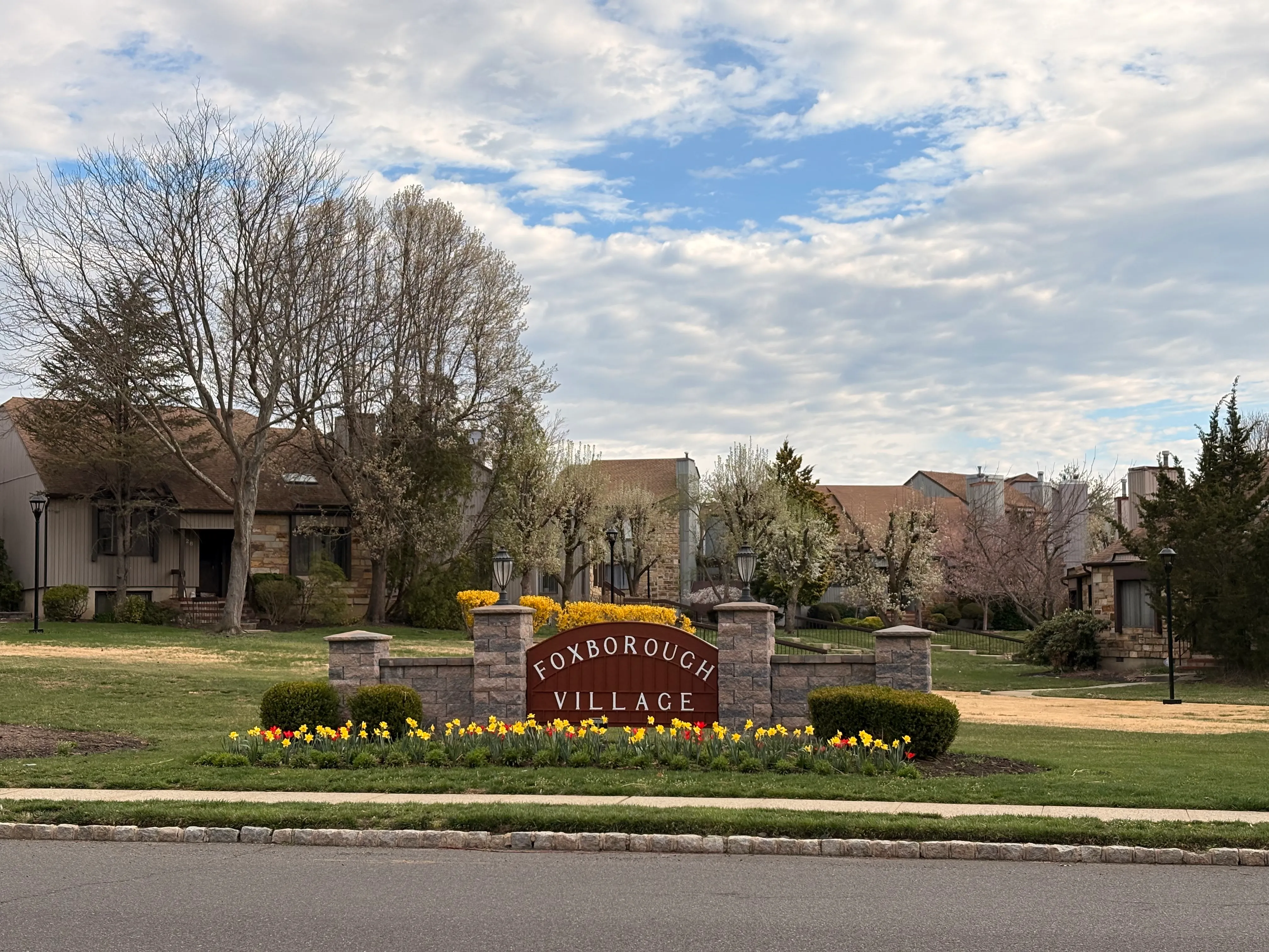 The Foxborough Village stone entrance sign on Emerald Lane, framed by spring daffodils and flowering trees.